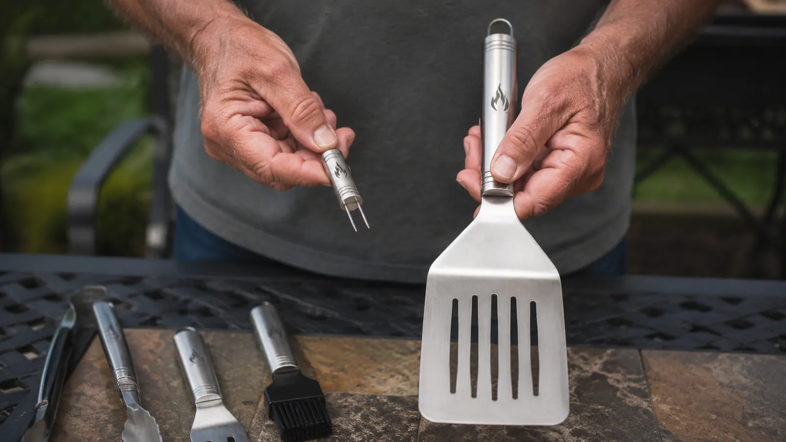 A man's hand holding a stainless steel corn holder skewer next to a stainless steel spatula on a patio table