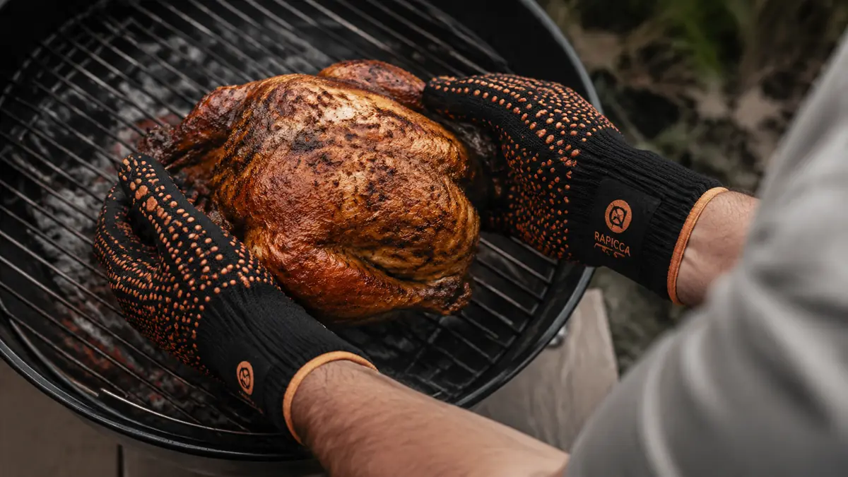 Two gloved hands cradling a whole cooked chicken from underneath while lifting it off the grill